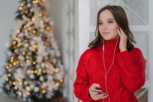 People, holidays and technology concept. Pretty woman uses mobile phone and earphones for listening music, stands at home against Christmas tree lights background with copy space for your text