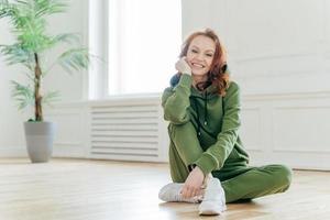 Healthy lifestyle concept. Beautiful slim young woman with red wavy hair, keeps hand under chin, smiles happily, dressed in sportswear, has flexible body, sits on floor. Female has home workout photo