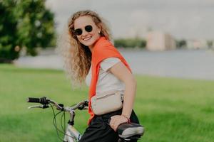 Satisfied curly female bicyclist enjoys spare time, travels on bike, stops to have little rest, wears sunglasses, casual summer outfit, poses among grass and trees, green blurred background.