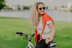 Active lifestyle concept. Positive young woman with curly light hair, rides bike on green lawn, wears shades, white t shirt and red sweater on shoulders, carries little bag, being in good mood