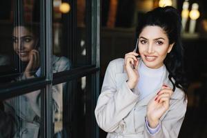 Pretty female with black hair having thin dark eyebrows, bright eyes with long eyelashes and full well-shaped lips wearing elegant clothing holding smartphone speaking with somebody and smiling