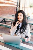 elegante y exitosa mujer independiente vestida formalmente usando una computadora portátil genérica para el trabajo remoto, descansando en un café al aire libre bebiendo un delicioso café con una expresión de ensueño. concepto de estilo de vida foto