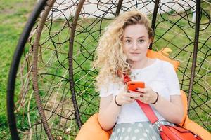 People and leisure concept. Beautiful curly female holds smart phone, types text message, looks at camera, has good rest in hanging chair outdoor, dressed in casual clothes, has appealing look photo