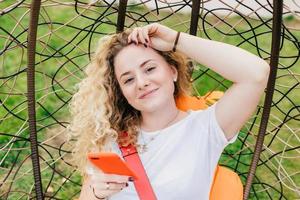 Beautiful delighted female blogger with curly light hair, uses smart phone for posting new posts on her web site, rests in hanging chair, uses wireless internet connection. People, recreation concept photo