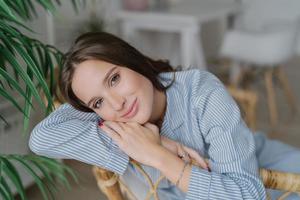 Image of good looking young female model with dark hair, sits on wooden chair, has relaxed facial expression, has charming smile, enjoys domestic calm atmosphere. Beauty and emotions concept photo