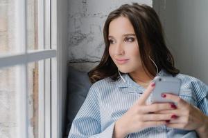 una mujer morena pensativa escucha un audiolibro, sostiene un teléfono inteligente moderno en las manos, disfruta de un sonido perfecto en los auriculares mira pensativamente a la ventana vestida con ropa informal. la mujer escucha música en la lista de reproducción foto