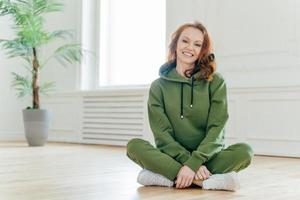 Horizontal shot of happy redhead sportswoman in green tracksuit, white sneakers, sits in lotus pose, has friendly smile on face, poses indoor. Female pilates instructor practices stretching exercises photo