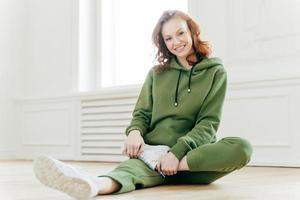 Photo of happy red haired woman in sweatsuit, white sneakers, takes rest after workout, sits on floor, stretches legs, has active healthy lifestyle. Smiling fitness girl does exercising. Flexibility