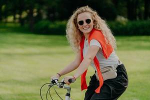 Photo of curly European woman keeps hands on handlebar of bicycle, travels outdoor in park, wears shades, casual t shirt and black trousers, smiles positively, expresses happiness. People, recreation