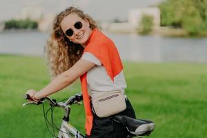 People and lifestyle concept. Cheerful young woman with crisp hair, wears casual clothes, shades, rides bike, has positive smile, poses in countryside, enjoys summer and fresh air during sunny day photo