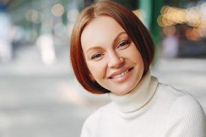 Image of attractive young woman with glad expression, looks directly at camera, has pleased look, wears turtleneck sweater, poses over blurred background, walks outside, expresses good emotions photo