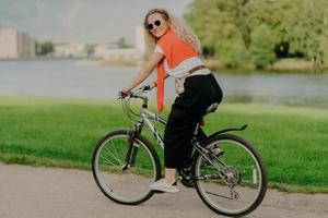 Pleased young woman enjoys new route on bicycle, rides among lake, green lawn and buildings far away into distance, wears summer shades, casual outfit, white sneakers, being in good physical shape photo