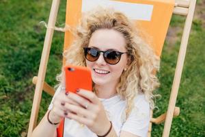 Positive European female with friendly smile, uses modern electronic device, happy to recieve message from boyfriend, rests outdoor during summer vacation. People, recreation, lifestyle concept photo