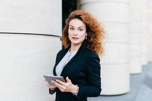 Adorable woman with curly hair wearing black suit and white blouse, holding tablet computer, typing messages or searching Internet while standing outdoor. Pretty businesswoman with modern device photo