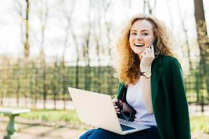 Attractive woman with trendy hairdo sitting in park communicating over her cell phone using modern laptop computer having broad smile while looking aside. People, technology, communication concept photo