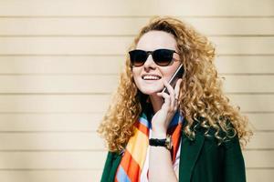 Young smiling office worker in sunglasses having blonde curly hair talking on cell phone during a break standing at street against beige background with copy space for your promotional content photo