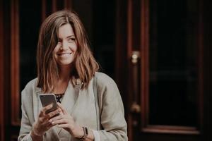 foto de una mujer guapa que busca información, usa un teléfono celular, se viste con una chaqueta elegante, posa contra el fondo de la puerta, se ve positivamente a un lado, se para al aire libre con un espacio en blanco para su texto