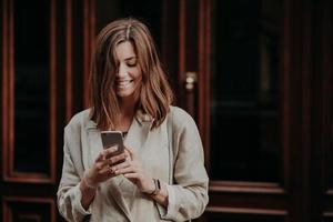 hermosa joven alegre recibe un mensaje de notificación, recibe una oferta publicitaria, tiene un corte de pelo moderno, se viste con ropa de moda, se para al aire libre, camina al trabajo, lee las últimas noticias en Internet foto