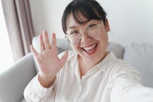 Young Asian woman using smartphone for online video conference call with friends waving hand making hello gesture photo