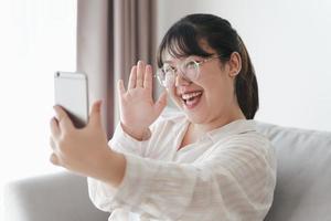 joven asiática que usa un teléfono inteligente para una videoconferencia en línea agitando la mano haciendo un gesto de saludo en el sofá de la sala de estar. foto
