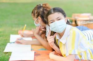 una niña medio tailandesa y su amiga usan una máscara antivirus mientras aprenden fuera de la escuela en un parque