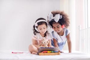 A cute Asian girl and an African kid friend are using a tablet to play games and have fun learning in the room photo