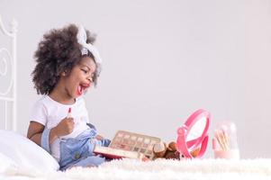 una linda niña africana se mira en el espejo y felizmente juega con el lápiz labial en sus labios en su dormitorio