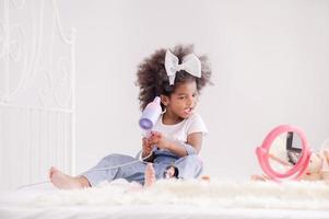 la linda niña africana felizmente usa un secador de pelo eléctrico en su dormitorio foto