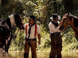 Old and young cowboys rest with their horses in the stream after they finish bathing photo