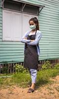 A woman wearing a mask stands with her arms folded close to the wall of a wooden house. photo