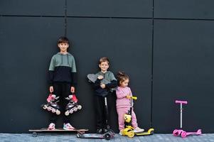 Three kids outdoor against black modern wall. Sports family spend free time outdoors with scooters and skates. photo