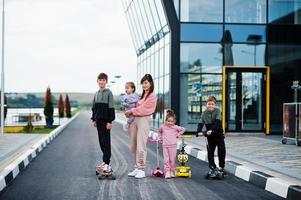 Young stylish mother with four kids outdoor. Sports family spend free time outdoors with scooters and skates. photo