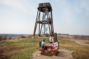 Mother with children sit at wooden table against observation tower. photo