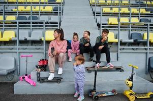 joven madre elegante con cuatro hijos sentados en el podio deportivo en el estadio, come manzana y bebe agua. la familia pasa tiempo libre al aire libre con scooters y patines. foto