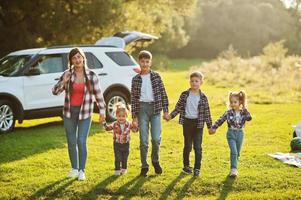 familia pasando tiempo juntos. madre con cuatro hijos de pie y cogidos de la mano contra un coche todoterreno blanco. foto