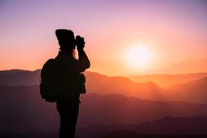 joven fotógrafa hipster tomando fotos con puesta de sol en el fondo natural de la montaña.