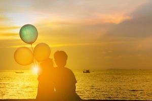 Silhouette of brother and sister sitting on the beach watching sunset with balloon in hand. photo