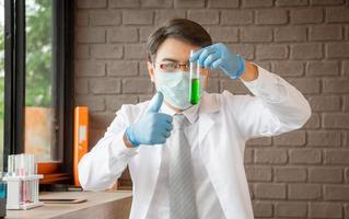 médico que analiza tubos de ensayo médicos que examinan el vaso de precipitados con líquido verde, científico que da el pulgar hacia arriba como señal de éxito en un laboratorio, concepto covid-19 foto