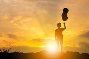 silueta de niño parado solo viendo la puesta de sol y sosteniendo un globo en la mano. foto