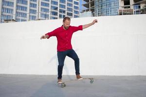 Young bearded man riding on skateboard, hipster with longboard in red shirt and blue jeans urban background photo