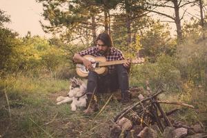 guitarrista folclórico o rural en el bosque con perro, hombre en el bosque, fogata y fondo de puesta de sol foto