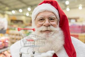 Santa Claus doing grocery shopping at the supermarket, he is showing a mini cart, Christmas and shopping concept.