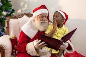 Santa Claus reading a storybook to a child on his lap. Education, beliefs and legends concept. Child delighted with what he learns. End of the year. Enchanted Fable. African boy. photo