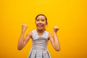 Young beautiful Asian little girl happily rejoicing saying yes. Success and achievement concept. Cheerful pretty Japanese kid on yellow background. photo