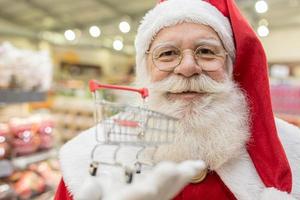 Santa Claus doing grocery shopping at the supermarket, he is showing a mini cart, Christmas and shopping concept.