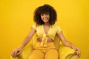 Happy laughing American African woman with her curly hair on yellow background. Laughing curly woman in sweater touching her hair and looking at the camera. photo