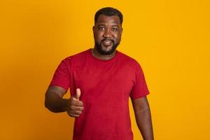 Positive dark skinned male has joyful expression and broad smile with white teeth, being in good mood, thumbs up, ok, okay isolated over yellow studio wall with copy space. Brazilian middle aged guy. photo