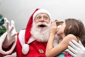 Little girl whispering in Santa's ear. Telling a secret. Revealing the gift you would like to win. Talking about how she behaved during the year. photo