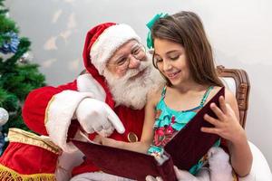 santa claus leyendo un libro de cuentos a un niño en su regazo. concepto de educación, creencias y leyendas. niño encantado con lo que aprende. fin de año. fábula encantada. foto