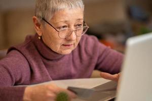 a senior woman with gray hair sits at a laptop with a credit card in her hands, makes online purchases. Online shopping concept photo